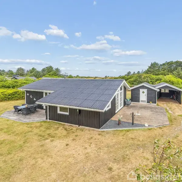 Et sort sommerhus står stille på en stor græsplæne med en terrasse og udendørs møbler. Omkring huset er der grønne buske og træer under en klar blå himmel.