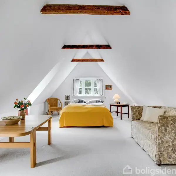 Et lyst loftværelse med synlige træbjælker. En seng med gul sengetæppe står i midten, omgivet af en bord med blomster, en sofa og en stol.