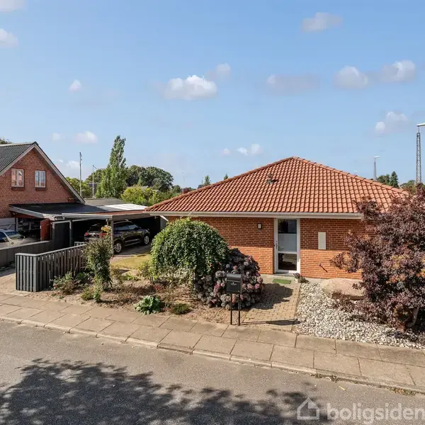 Et rødt murstenshus med et tegltag står stille i et villakvarter. Foran huset er der en have med træer og buske. En bil og en carport ses ved siden af.