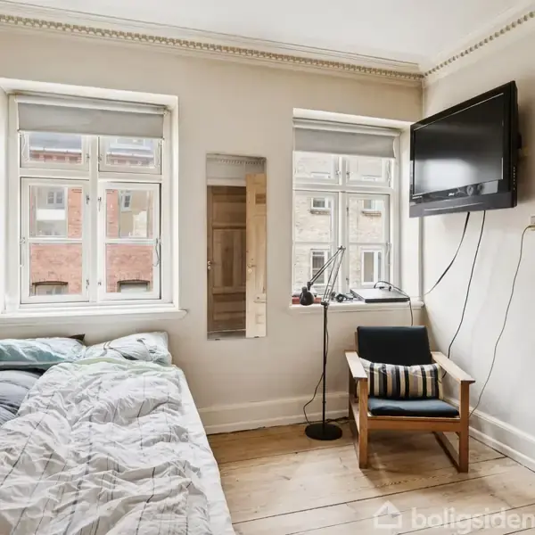 En seng med sengetøj står op ad en væg under to vinduer. Ved siden af hænger et fjernsyn, og en stol med pude står nær et bord med en lampe.