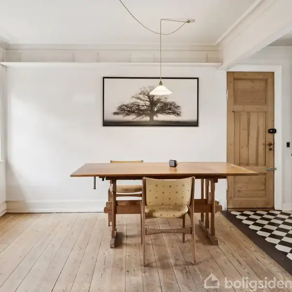 Et spisebord med to stole står på et trægulv i et lyst rum. På væggen hænger et billede af et træ. Ved siden af er et køleskab og en trædør.