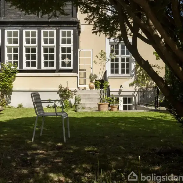 En metalstol står på en græsplæne foran et klassisk gulmalet hus med sorte vinduesrammer. Trappen ved huset er flankeret af blomsterkrukker og plantebeplantning.