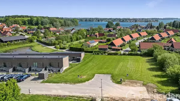 Et boligområde med moderne rækkehuse ligger ved en vej omgivet af græsplæner. I baggrunden ses flere huse med røde tage og en sø omkranset af skov.