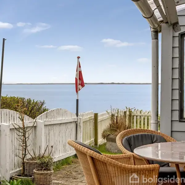 Et bord med kurvestole står på en terrasse. Udsigt til havet med en dansk flagstang i baggrunden. Træhegn og beplantning omkranser området.