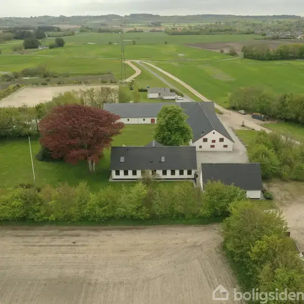 En gård omgivet af grønne marker og træer. Bygningerne ligger omkring en gårdsplads. Store træer markerer kanten af ejendommen. Landskaber strækker sig i baggrunden under en overskyet himmel.