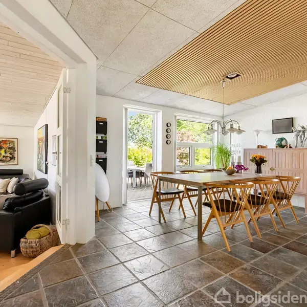 Et moderne spisestueområde med ti træstole omkring et bord. Det åbner ud til en lys stue og køkken med store vinduer, der slipper naturligt lys ind. Skiferfliser dækker gulvet.