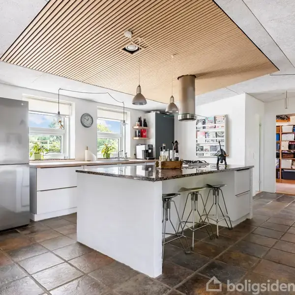 Køkken med moderne køleskab og central ø, der har barstole. Bordet er fyldt med kogebøger og køkkenredskaber, omgivet af skabe og store vinduer, der lukker naturligt lys ind.