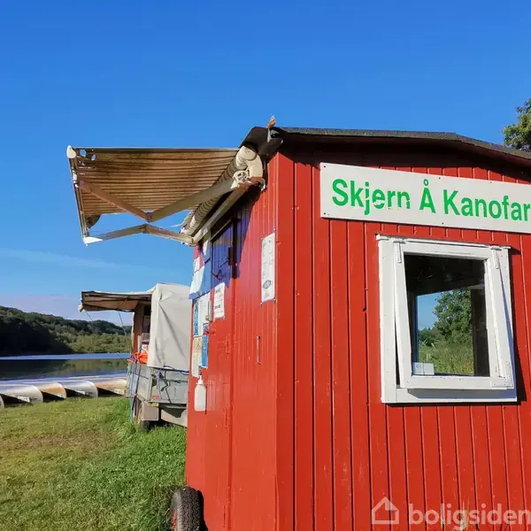 En rød træbygning fungerer som kiosk ved siden af en flod. Skiltet på bygningen lyder "Skjern Å Kanofart". Flere kanoer ligger ved flodbredden under en klar blå himmel.