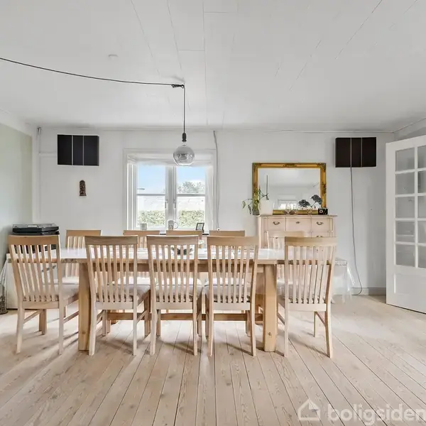 Spisebord med otte stole står i lys spisestue med trægulv. En lampe hænger over bordet. En kommode og et spejl står i baggrunden nær et åbent vindue.