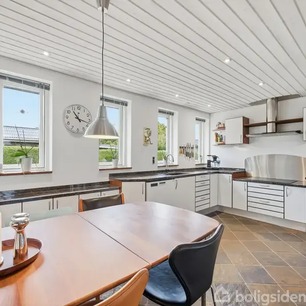 Et køkken med hvidt træloft og flisegulv har et spisebord i forgrunden. Der er tre vinduer, vægur, og moderne køkkenelementer. Indretningen er lys og funktionel.