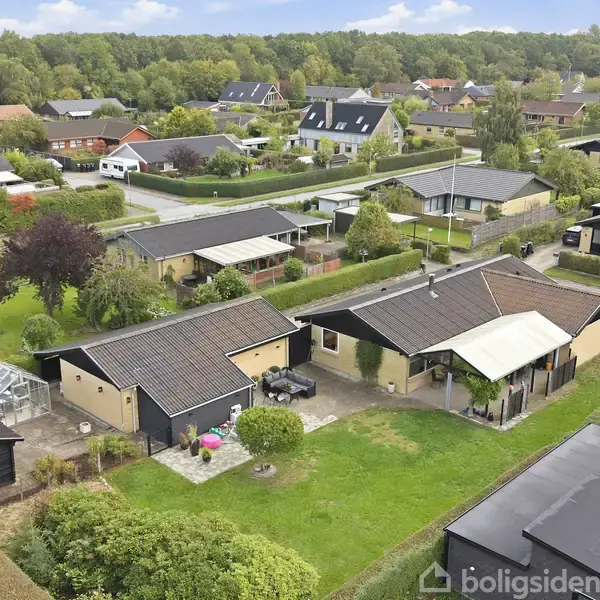 En villa med sort tag og carport er omgivet af grønne haver og buske. Huset ligger i et parcelhuskvarter med flere nærliggende boliger og træer i baggrunden.