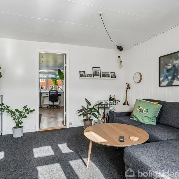 En stue med en grå sofa, et rundt træbord og et tæppe. Vægdekorationer og planter skaber hygge. Et åbent dørkar til et andet rum med kontor ses i baggrunden.
