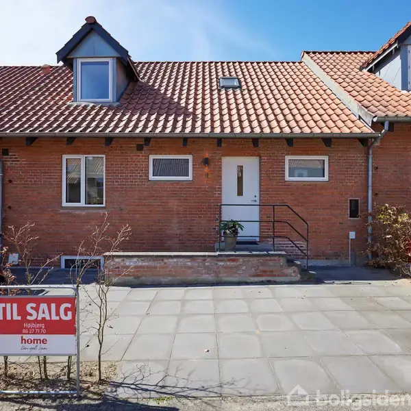Rækkehus vises med rød murstensfacade og tegltag i en forstad. Skilt foran huset viser teksten "TIL SALG home mægler Nanna Revisen." Detaljeret indkørsel og postkasse i forgrunden.