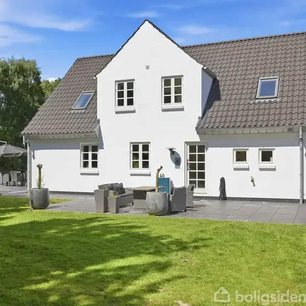 Hvidt hus med gråt skråtag står i en grøn have. Udendørs terrasse med havemøbler og parasol. Træer omgiver huset i baggrunden.