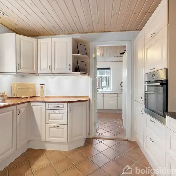 Et lyst køkken med hvide skabe og træbordplade. En vask står under et vindue, mens en ovn er indbygget til højre. Fliser dækker gulvet, og gangen fører til et tilstødende rum.