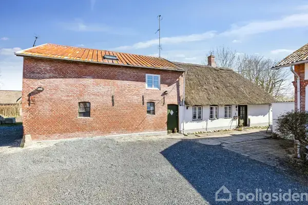 Et gammelt landhus med en rød murstensfacade står stille ved siden af et bindingsværkshus med stråtag, omgivet af en grusbelagt gårdsplads og klar blå himmel.