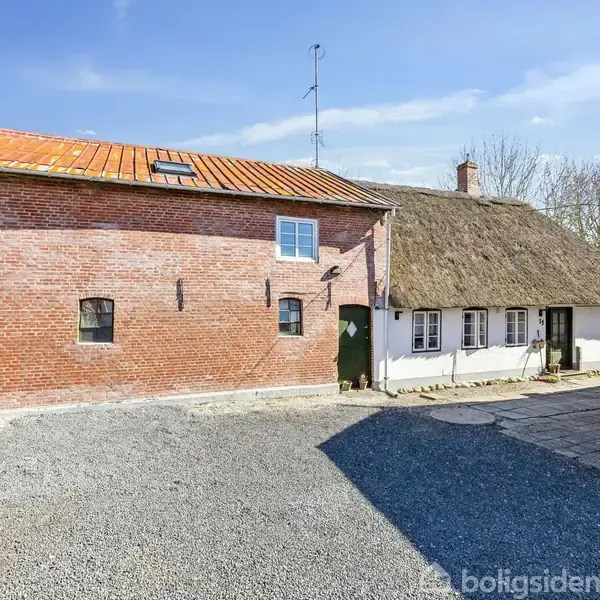 En rød murstensbygning med et skråt tag grænser op til et bindingsværkshus med stråtag. Begge bygninger er placeret på en grusbelagt gårdsplads under en klar blå himmel.
