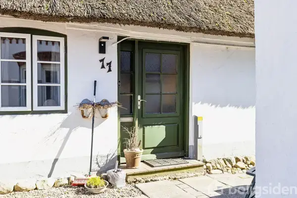 En dør med grøn ramme står stille mellem hvidkalkede vægge, beliggende i en gårdhave med brosten. Ved siden af er to plantepotter og husskiltet "11" i sort metal placeret.