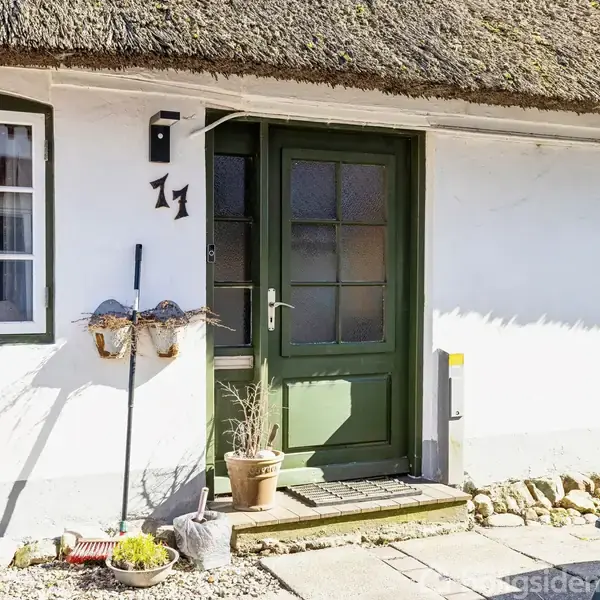 Dør i traditionelt hus med grøn ramme, åbner mod en lys gård. Potteplanter står ved siden af dørtrinnet. Husets facade er hvid med små vinduer. Nummer 11 er over døren.