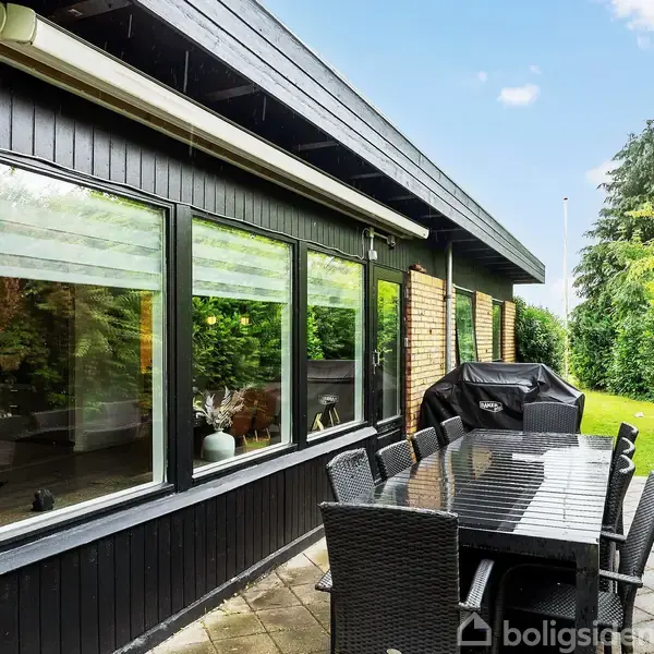 Sort havebord med stole står på terrasse ved moderne hus med store vinduer. En grill er placeret ved siden af bordet. Omgivet af frodig grøn have og hække.