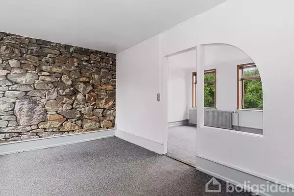 Værelse med en stenvæg til venstre, hvidmalede vægge og en grå gulvbelægning, åbner op til et andet rum med store vinduer og udsigt til grønne træer.