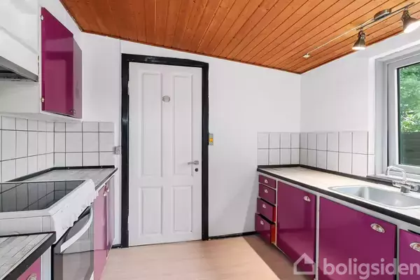 Køkken med lilla skabe og hvid dør, hvide fliser på væggene og træloft. Et vindue med udsigt til grønne omgivelser til venstre for vasken.