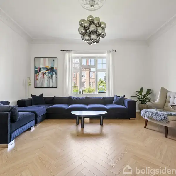 En stor, mørkblå sofa står i en stue med trægulv, flankeret af et moderne sofabord. Et vindue i baggrunden giver udsigt til bygninger udenfor, mens kunst pryder væggene. En stor lampe hænger ned fra loftet, og en stor lænestol står ved siden af et træbord til højre.