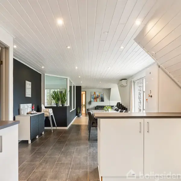 Åbent køkken med hvide skabe og gråt gulv, lysindfald fra vinduer til højre. I baggrunden ses spisestue med sort bord og stole. Moderne og minimalistisk indretning.