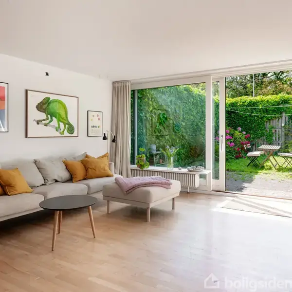 En stue har en grå sofa med gule puder, stående foran en væg med kunstbilleder. Store vinduer åbner op til en frodig have med blomster og et lille bord.