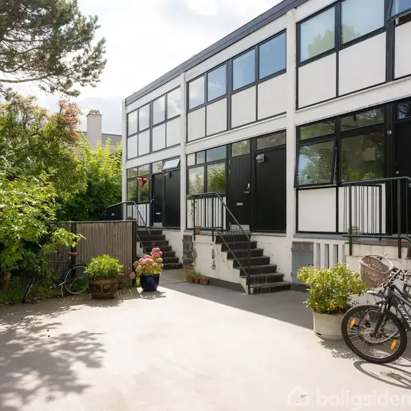 To etager rækkehus med sorte døre og vinduer; trapper foran indgange. Cykler og blomsterkrukker står i gårdlignende område omkranset af grøn vegetation. Ingen synlig tekst i billedet.