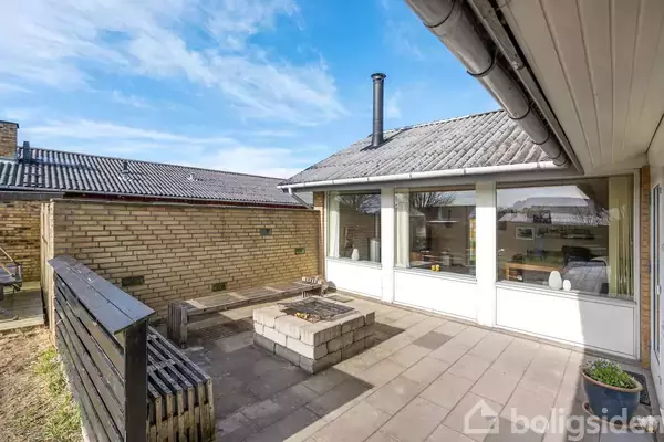 Terrassen har en muret pejs i midten, omgivet af flisebelægning og murstensbygninger. Store vinduer åbner ind til husets indre, hvor man kan se indretningsdetaljer.