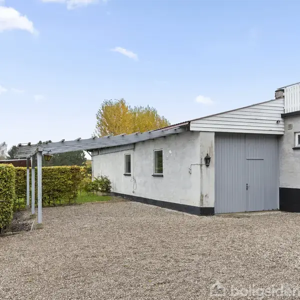En hvid bygning med en grå garageport står stille ved siden af en grusbelagt indkørsel. En velplejet hæk og et gammeldags gadelys er synlige i en landlig have.
