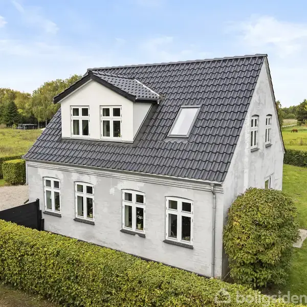Et hvidt murstenshus med sort tegltag står omgivet af en velholdt græsplæne og hække. Der er åbne marker og træer i baggrunden under en klar, blå himmel.