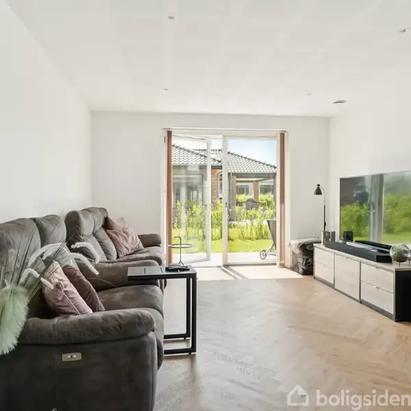 En stue med en grå sofa fyldt med puder placeret til venstre, en moderne fladskærms-TV til højre på et hvidt TV-bord, og store glasdøre fører ud til en have.