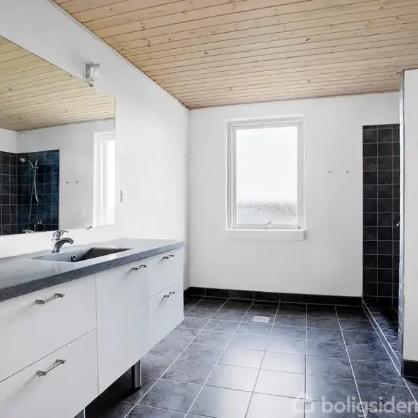 Badeværelse med hvidt skabsmodul og sort vask, spejl ovenfor. Bruseområde til højre med mørke fliser. Trægulv og træloft. Et vindue giver naturligt lys.