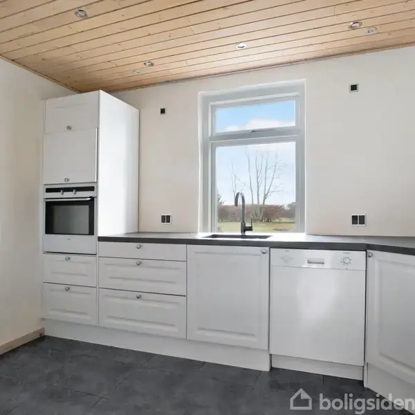 Et lyst køkken med hvide skabe, en indbygget ovn og opvaskemaskine. et vindue over vasken giver udsigt til en solrig have. Træloft og grå flisegulv skaber en moderne atmosfære.