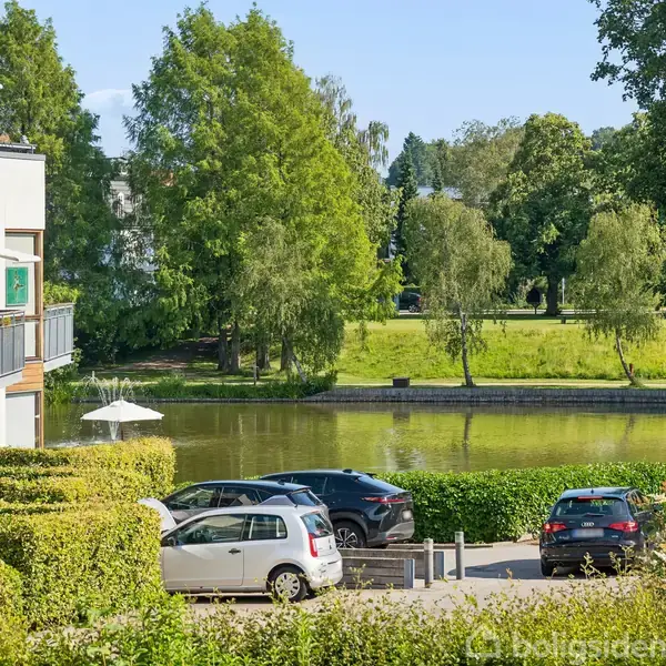 En moderne bygning med altaner står til venstre. Biler holder parkeret foran bygningen. En flod flyder bagved med velplejede grønne områder og træer i baggrunden.