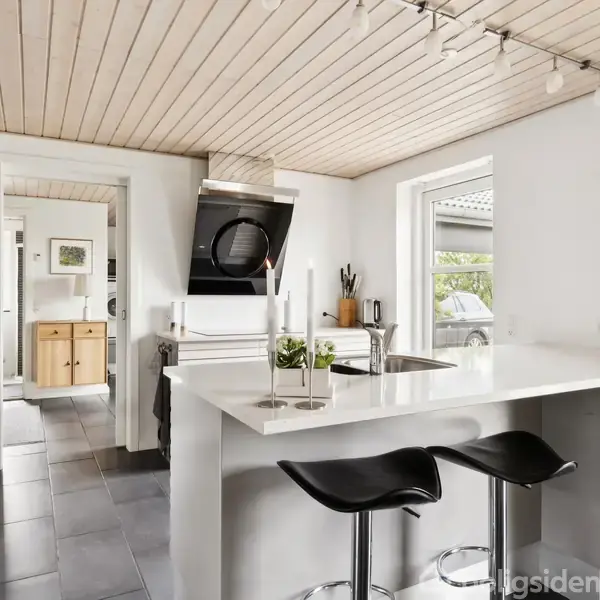 Køkken med hvid køkkenø, to sorte barstole. Bordplade har planter og stearinlys. Moderne emhætte over kogeplade. Lyse vægge og træloft. Stort vindue giver naturligt lys.