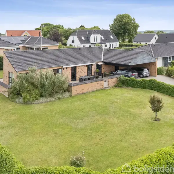 En moderne murstensvilla med sort tag ligger i en velplejet have med grøn plæne og omkransende hæk. I indkørslen holder to biler. Flere lignende huse ses i nabolaget.