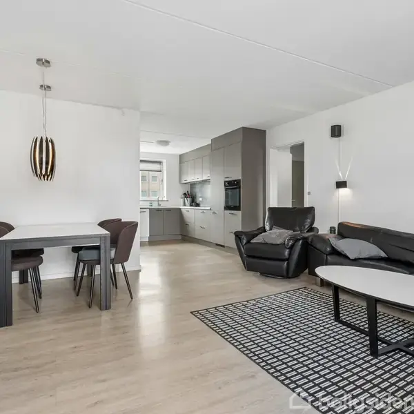 Sort læder sofa og lænestol står i en moderne stue med et sort-hvidt tæppe. Spisebord med stole og kunstlampe ses nær køkkenet. Billeder hænger på væggene.