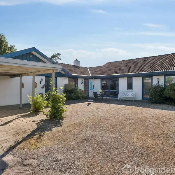 Et parcelhus står stille i en solrig gårdsplads med en carport til venstre. Huset har et tegltag og store vinduespartier, omgivet af buske og en stenbelagt indkørsel.