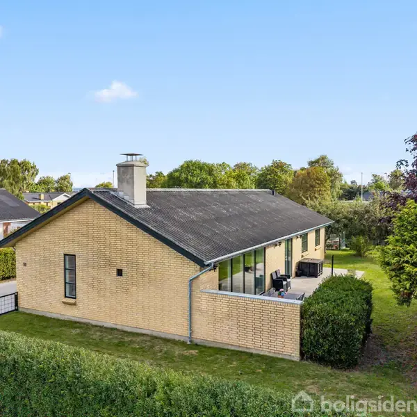 En gulstensvilla med sort tag ses fra siden, omgivet af velplejede hække og buske i en rolig, grøn have. Terrasse med havemøbler findes ved siden af huset.