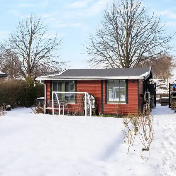 Et lille, rødt træhus står stille i en snedækket have omgivet af hække. Træerne uden blade ses i baggrunden, og himlen er let skyet.