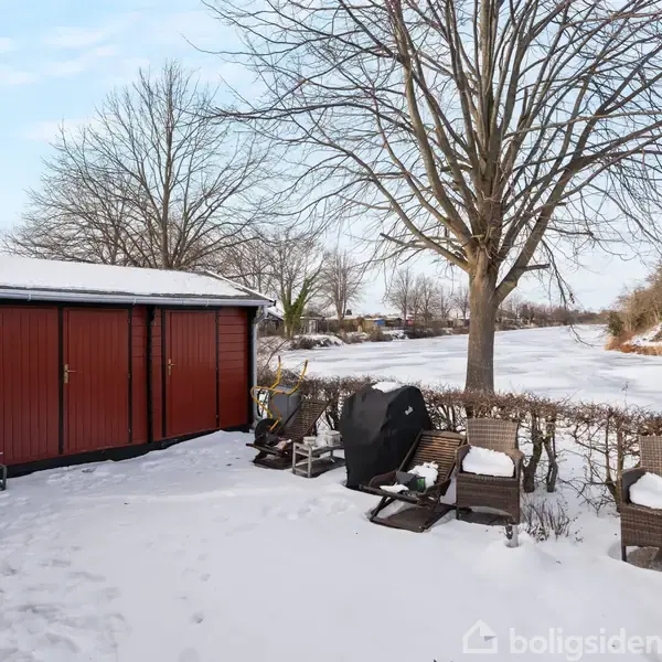 Et rødt skur står stille i en snedækket have med en dartskive på siden, omgivet af havemøbler og en snedækket hæk. I baggrunden ses træer og en frossen sø.