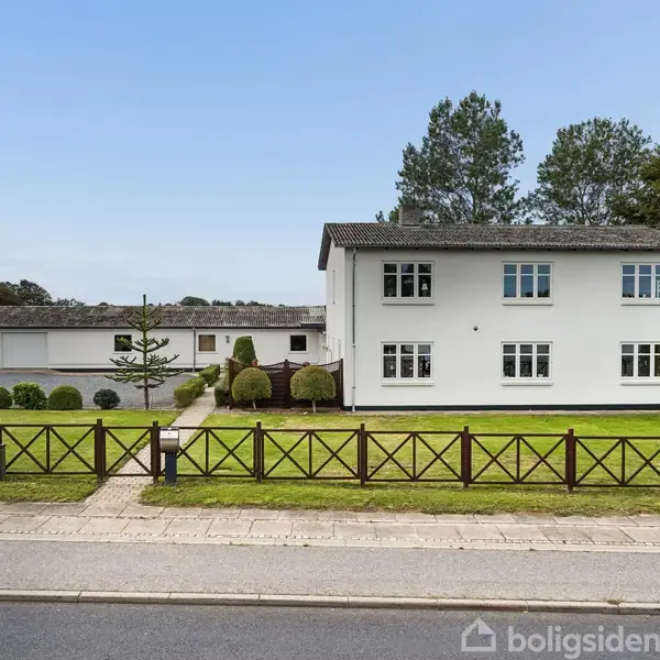 Huset har en hvid facade med en træterrasse på første etage. Terrassen har trappeadgang og udsigt over en lille have omkranset af træer og hække.