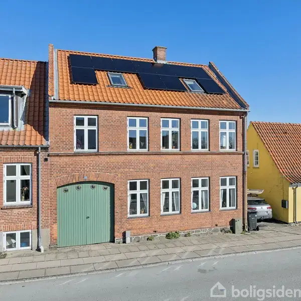 Rød murstenshus med solpaneler på taget står stille på en gade. Det har grønne porte og hvide vinduer. Ved siden af er et gult hus. Klar blå himmel.