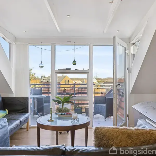 En lys stue med sorte sofaer, et rundt bord, og planter står mod en stor glasvæg. Bag ruderne ses en balkon med stole, udsigt til blå himmel og bygninger.
