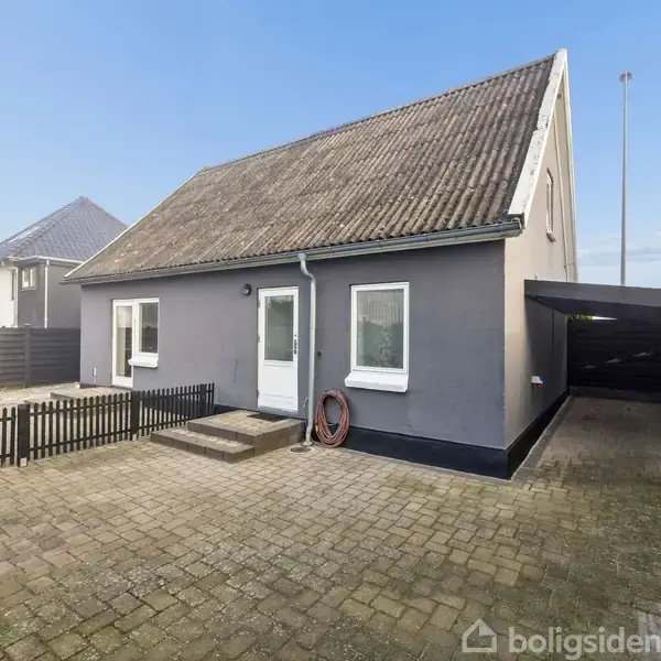 Gråt hus med skråt tag, omgivet af en hæk og en postkasse foran. Det står i et villakvarter med fortov. Der er en lille busk og stakit ved siden.