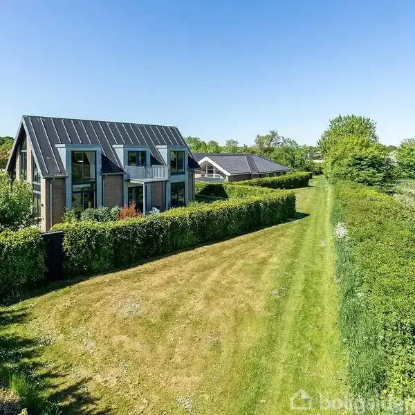 Et moderne hus med store vinduer står blandt grønne hække og en velplejet græsplæne. Baggrunden er omgivet af frodig natur og en klar blå himmel.