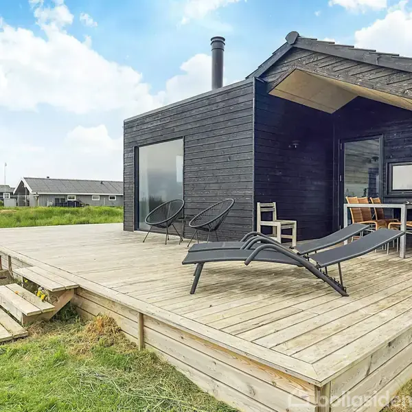 Et sort træhus står stille på en træterrasse i en grøn have. Terrassen har liggestole, stole og et bord, hvilket skaber en afslappet atmosfære under blå himmel.